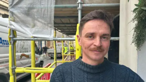 A man with a in a navy sweater, with a moustache and light brown hair smiles at the camera with yellow scaffolding and a street behind him. 