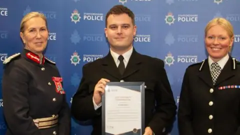 Staffordshire Police Three police officers standing in front of a blue background with the staffordshire police logo printed all over it. Two women have blonde hair tied back and are wearing black police jackets with large silver buttons. On the left, the woman has several red badges on her breast and badges on her shoulders. The other woman has a black and white checked tie. There is a man in the middle holding a certificate. he is wearing a black jacket, white shirt and black tie