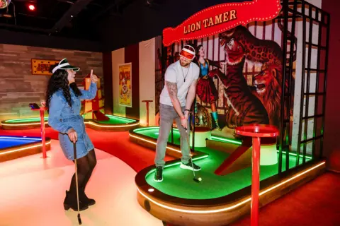 Carnival Golf and Games A couple are pictured playing mini-golf. A woman on the left with long dark wavy hair, wears a denim dress, boots and a visor on her head. She is holding a golf club. A man is standing on the putting green in a red visor, white tee-shirt and jeans. He is in front of a sign saying Lion Tamer