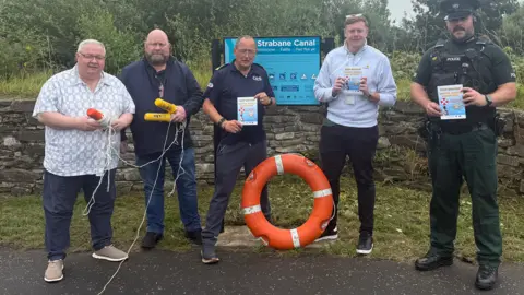 PSNI Five men, including on the far right a police officer in unifrom, stand in front of a sign saying Strabane Canal - behind the sign sits the canal itself. Each of the men are holding life saving equipoment