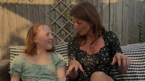 Mat Davina McCall sitting down on garden furniture outside talking to a female child in a green floral dress.