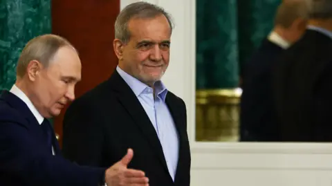 Reuters Vladimir Putin, seen in profile, gestures towards his Iranian counterpart, Masoud Pezeshkian, who is walking slightly ahead of him. Both men are wearing dark suits. Putin has a white shirt and dark tie, while Pezeshkian wears a blue open-necked shirt.