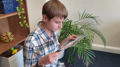 The image shows Jack, wearing a short-sleeved checked shirt, sitting down looking at a photograph of a spider on a daisy.