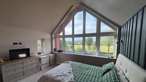 Luke Deal/BBC A newly redecorated and renovated master bedroom inside a home. There is a bed with a small TV opposite on a chest of drawers. To the rear of the room is a large window that overlooks countryside. 