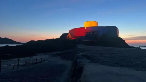 Un castillo gris con forma de taza y platillo, con una luz roja y naranja que brilla en uno de sus lados.