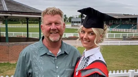 A red haired man in a green open collared shirt, stood next to him is his daughter in a graduation cap and gown with a police uniform underneath. they are both smiling at the camera.