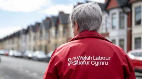 A woman wearing a red Welsh Labour jacket, with the party's logo emblazoned in white. The woman has grey hair and can be seen walking up a road with terraced housing, which is out of focus. Her back is to the camera.