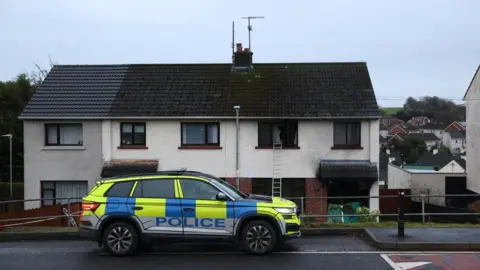 A yellow and blue police car is parked outside a row of terrace houses. The house on the right has burnt out windows and a ladder leading to the upstairs left window.