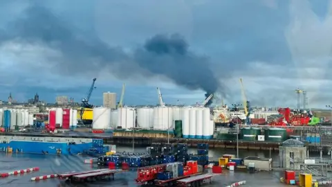 A large plume of dark smoke drifting across the blue skyline in Aberdeen as seen from the harbour area.