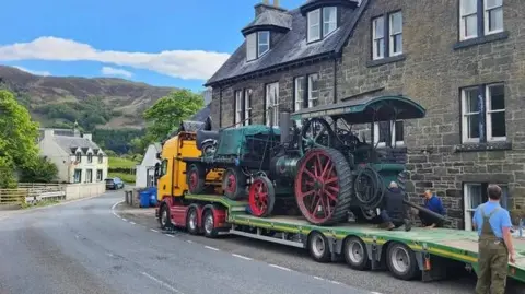 Rachel Rennie Two traction engines on the back of a tow truck on a rural road. There are stone houses beside the truck and hills in the distance.