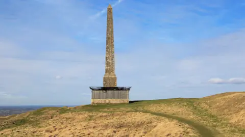 A tall stone obelisk on a hillside. At the base of the structure is wooden fences and netting.