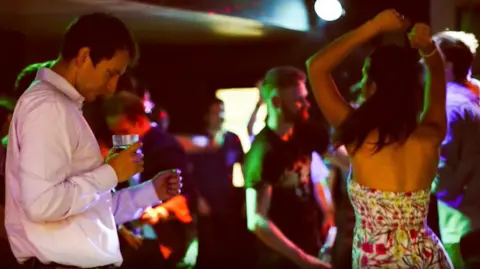 A man and a woman dancing in a nightclub setting, with multicoloured disco lights. The man has short brown hair is wearing a pink shirt and holds a glass, and the woman has long brown hair is wearing a strapless white dress, patterned with pink and green flowers.