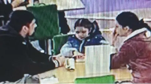 A couple with dark hair sit at a table with their daughter, who also has dark hair and wears a blue and white jacket. The father wears a dark jacket and the mother wears a red jacket with white lining.