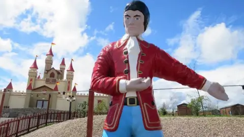 A giant plastic model of the fictional character Gulliver - a man in a white scarf, red jacket with gold trim, and blue trousers - stands at the entrance to a theme park. He appears to be gesturing people to walk to his left. Behind him is a small replica castle, with eight towers topped by red turrets, bearing yellow and red flags.