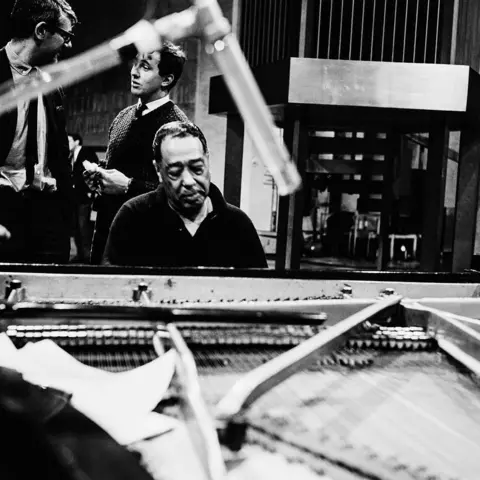 Getty Images A black and white image of Ellington at the piano. He is wearing a dark shirt, with two men seen standing behind him talking