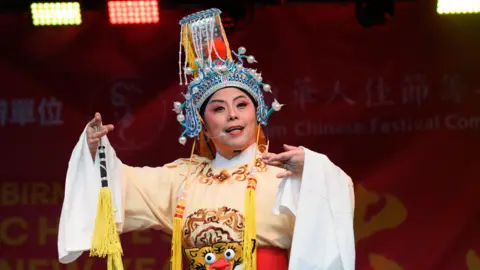 A performer in a traditional Chinese opera costume stands on a stage with bright lighting overhead. The outfit includes a cream robe decorated with patterns. The backdrop is red with festival branding faintly visible behind the performer. 
