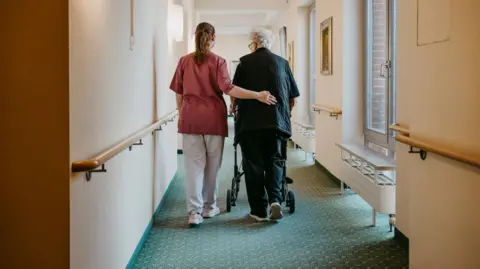 Inside a care home where a member of staff is gently guiding an older resident down a corridor with green carpets and beige walls.