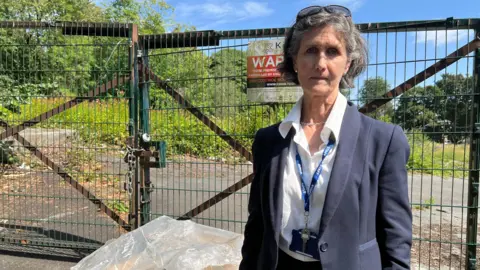 Aisha Iqbal/BBC A middle-aged woman in a dark blue suit jacket and white shirt. She stands in front of a gated-off area with an abandoned site behind it with overgrown greenery and paths visible. There is also a security/warning sign posted on the gate.