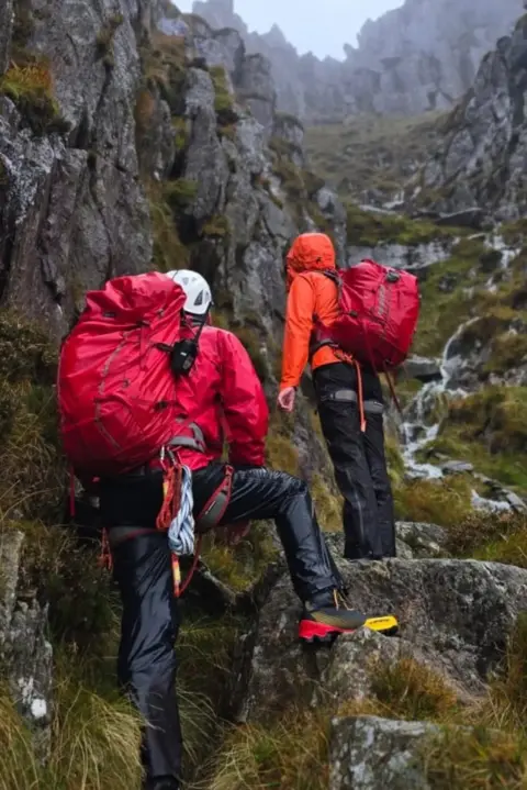 Ogwen Valley Mountain Rescue Organisation Two mountain rescuers in high visibility red clothing and backpacks head up a mountain pass in the rain. 