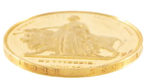 Rogers Jones and Co a close up of the gold coin looking from below with the latin words anno regni on the round edge. the coin face shows a lion walking submissively beside a woman in a flowing queen victorian style gown and holding a rod over the lion's head. 