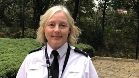 Insp Laura Stevenson is standing in the outside area of a police investigation centre. It has fencing and hedges in the background. The inspector is wearing her police uniform with tie and is smiling.