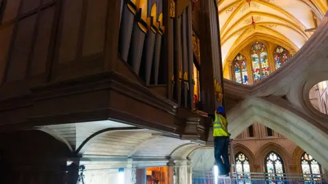 Wells Cathedral A wide shot of a man stood on a ladder working to remove organ pipes. The organ is high above the ground and the cathedral's stone walls and stained glass windows can be seen in the background.