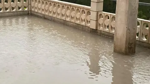 Diana Whitwell A flooded terrace in a country house. Water covers the terrace surrounded by decorative railings.