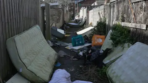 An alleyway littered with delivery driver bags, dirty mattresses, sofas and litter.