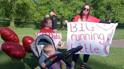 A lady with long brown hair and sunglasses holds a white sign as she wears a red top, black trousers and sandals. Beside her is a young girl holding a white sign and wearing a red top, and a small boy in a grey pram.