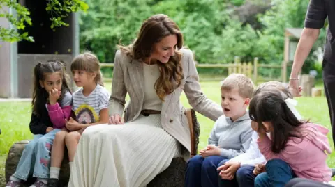The princess sits on a log with two children on her left and three to her right. She has her hand the back of one boy who has short fair hair and is dressed in a grey top and navy blue jeans and trainers