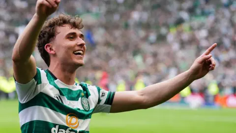 PA Media Callum is in a green-and-white hooped Celtic football shirt raises both index fingers while facing a crowded stadium, with blurred spectators filling the background.
