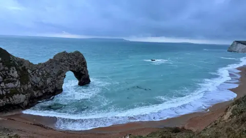 The arch of Durdle Door seen from a high vantage point with the beach in the foreground and a choppy-looking sea in the background beneath cloudy skies. 
