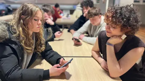 Nina McNair and Evi Barzel sitting at a table while one looks at a phone.