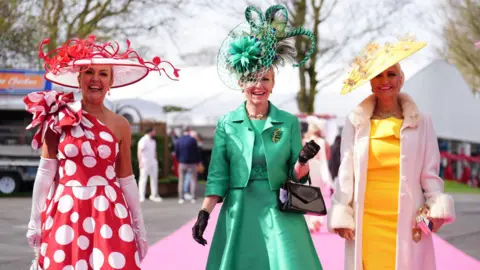Three women walking in on a pink carpet. On eis dressed in a red and white polka dot dress and hat, one is wearing a light green dress and hat and the third is wearing a yellow dress and hat and white coat