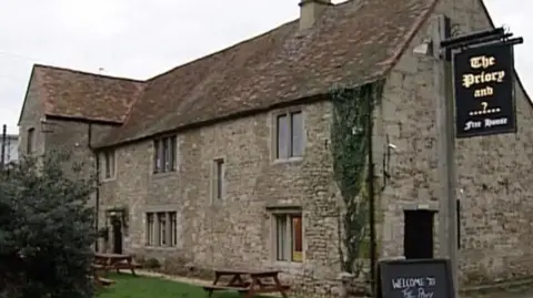 An old two storey limestone public house with a tiled roof and some picnic benches outside