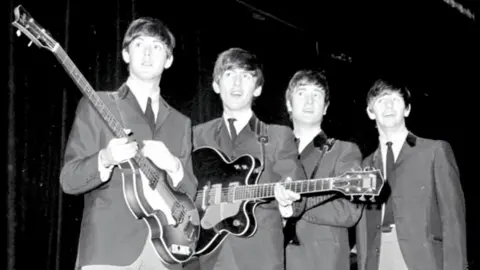 PA Media A black and white photograph of all four members of The Beatles holding guitars backstage on their 1963 tour.