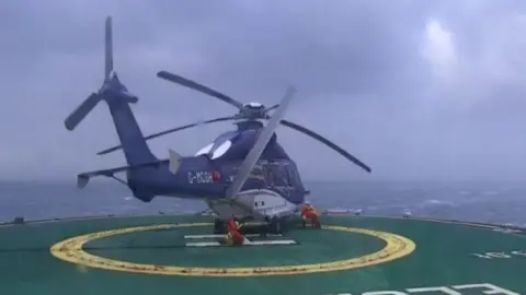 The blue and white helicopter is slightly tilted to one side on a green and yellow helipad. One of the rotor blades is broken. In the background is a stormy sea and clouds.