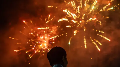 A man with short dark hair watches orange and yellow fireworks 