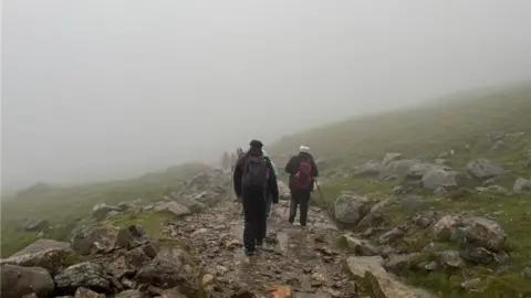 Worcestershire Acute Hospitals NHS Trust Several people are hiking on a rocky mountain path. There is dense fog all around them. They are wearing hiking gear and have hiking poles. You can make out green grass and grey rocks either side of the path