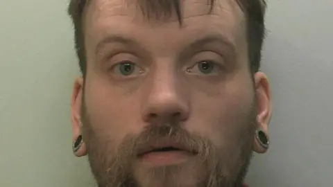 The image shows a close up of a mug shot of a man. He is staring at the camera. 
