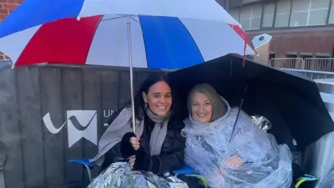 Michelle Power and Karen Dunkley, two women, one in a black coat and grey scarf is grinning while holding a blue, red and white umbrella and the other woman in wearing a translucent poncho and holding a black umbrella