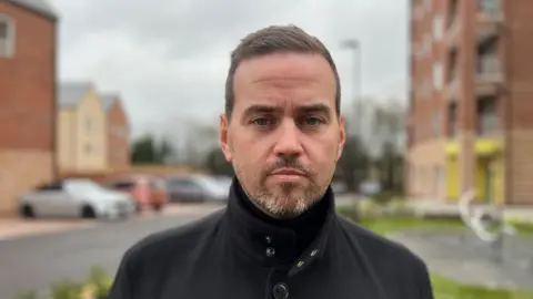 A man in his 30s with a short beard wearing a fitted black jacket. He is standing in a residential street with residential flats in the out-of-focus background.