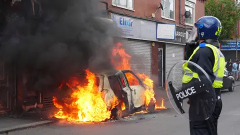 PA Car burning on a high street while a police officer looks on.