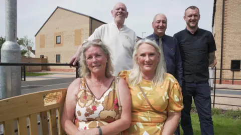Noreen’s daughters, Dawn Radford and Lynne Daffern, sit on the bench by the plaque. They have patterned summer dresses on. Son, Alan Turner is stood behind with deputy leader Steve Evans, and Morro Partnerships Contracts Manager, Matt Jenkins. 