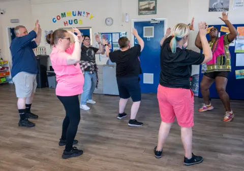 TeesDance A community dance class. Five people are facing a dance instructor and copying a dance move where their hands are up in the air.