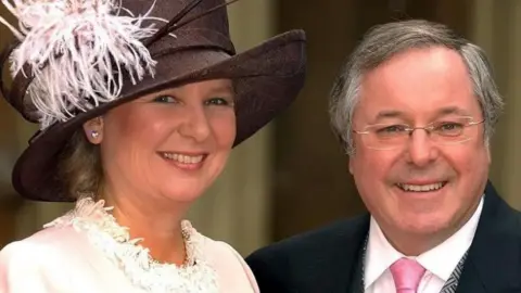 Kathryn Apanowicz is smiling and wearing a cream dress and brown formal hat with cream feather detail. Richard Whiteley is also smiling and is wearing a black three-piece suit with white shirt and pink tie. 