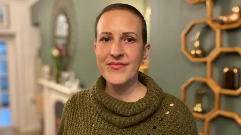 BBC Rachel Davies looking at the camera. She is wearing a green jumper and has her hair tied back, and is standing in her living room.