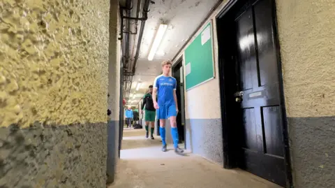 Players walking through the corridor of the changing rooms. A player at the front has brown curly hair and is wearing a blue football kit. The ceiling of the changing rooms has flaking paint.