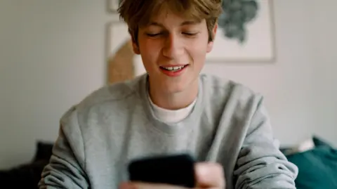 Getty A teenage boy with short light brown hair sits indoors, holding and looking at a black smartphone. He wears a light grey sweatshirt over a white shirt. The blurred background features a painting on the wall and a blue sofa behind him.