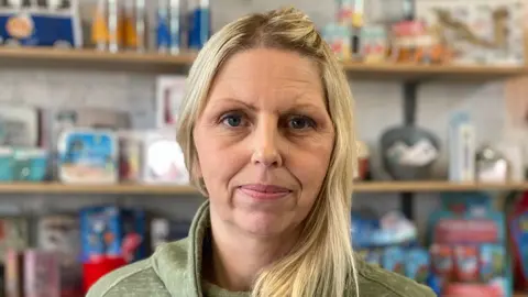 Harry Parkhill / BBC News Sophie Kirk with blond hair wearing a green top standing in front of a row of shelves in her shop